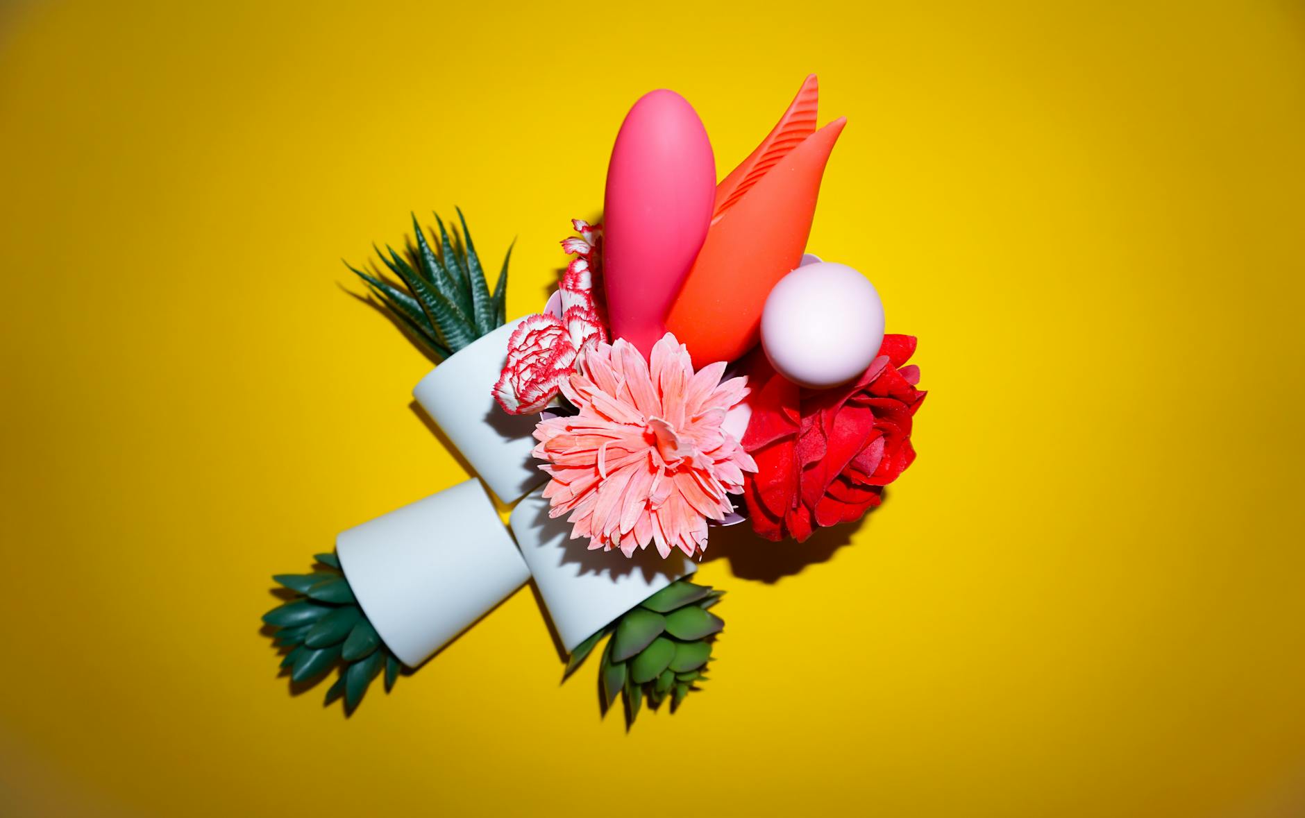 Colorful vibrators with flowers in a holographic gift bag, set against a bold yellow background.